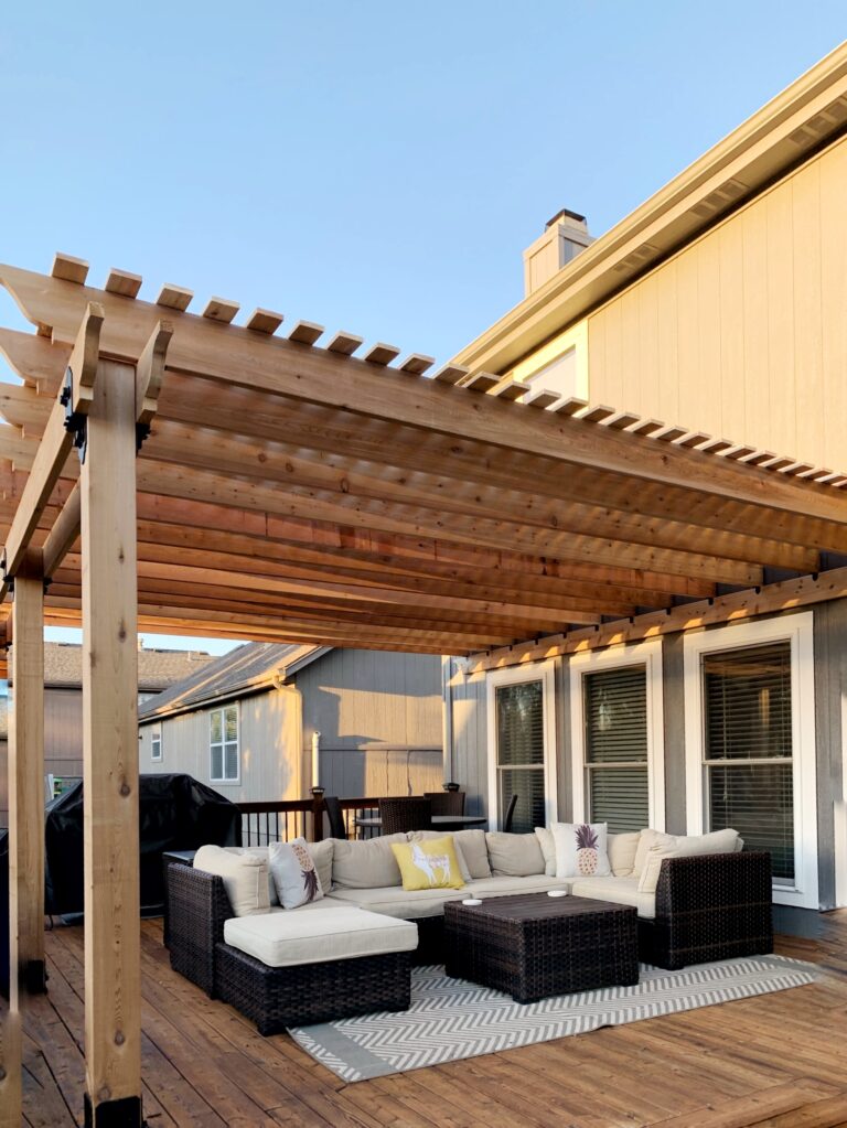 Cozy Outdoor Seating Area Under a Wooden Pergola, Featuring a Sectional Sofa and Coffee Table, Ideal for Relaxation or Gatherings.